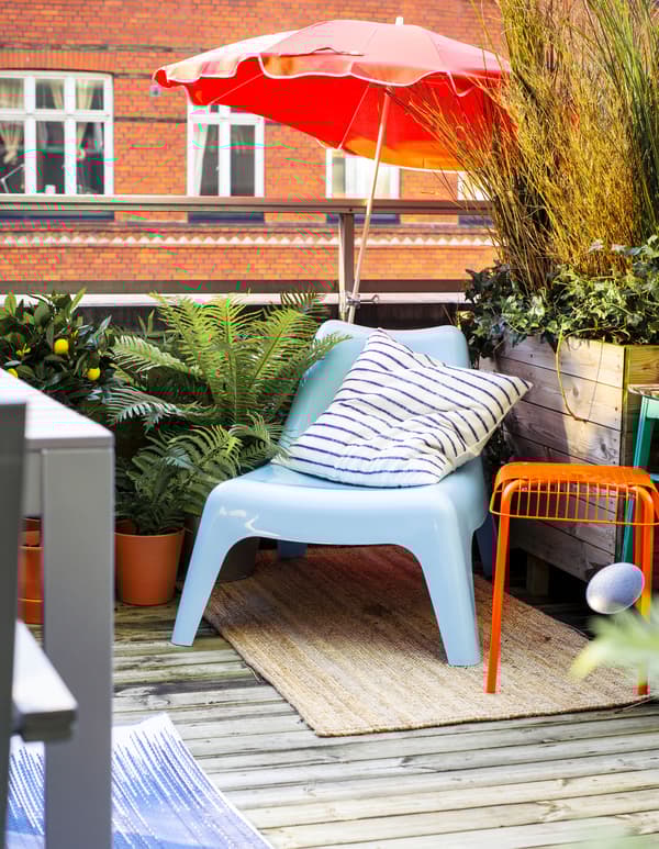 Fauteuil bleu dans le coin d’un balcon, entouré de plantes vertes, avec grand parasol.