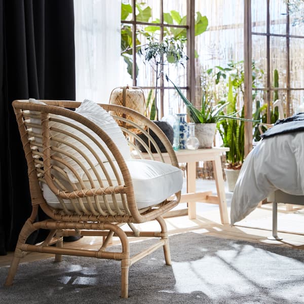 Fauteuil BUSKBO en rotin, coussins blancs, rideaux gris foncé et blancs, banc, plantes vertes et tapis gris 