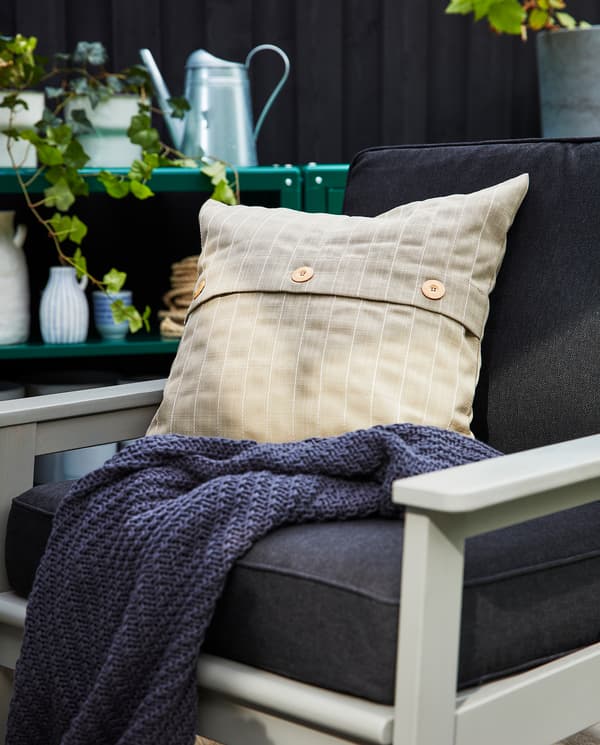 Fauteuil d’extérieur gris avec coussins d’assise et de dossier en gris foncé, coussin d’extérieur beige et plaid gris foncé.