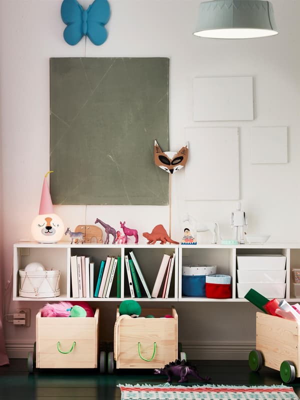 Fehér, falra szerelt polcos elem. Játékok, könyvek, kosarak és asztali lámpa a polcokon és a tetején.