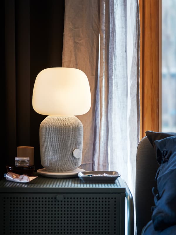 Fehér SYMFONISK lámpa és WiFi hangszóró áll egy zöld éjjeliszekrényen, a sötét szobában lámpa világít.