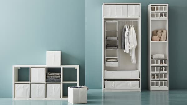 Fehér tárolórendszer nyitott polcokkal, textil dobozokkal és ruhafüggő szervezőkkel kék fal ellen.