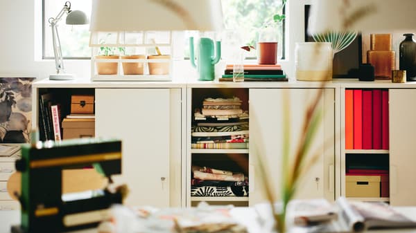 Fehér TROTTEN tolóajtós szekrények, az egyiknek a tetején egy élénkzöld ÄRTBUSKE váza/locsolókanna látható.