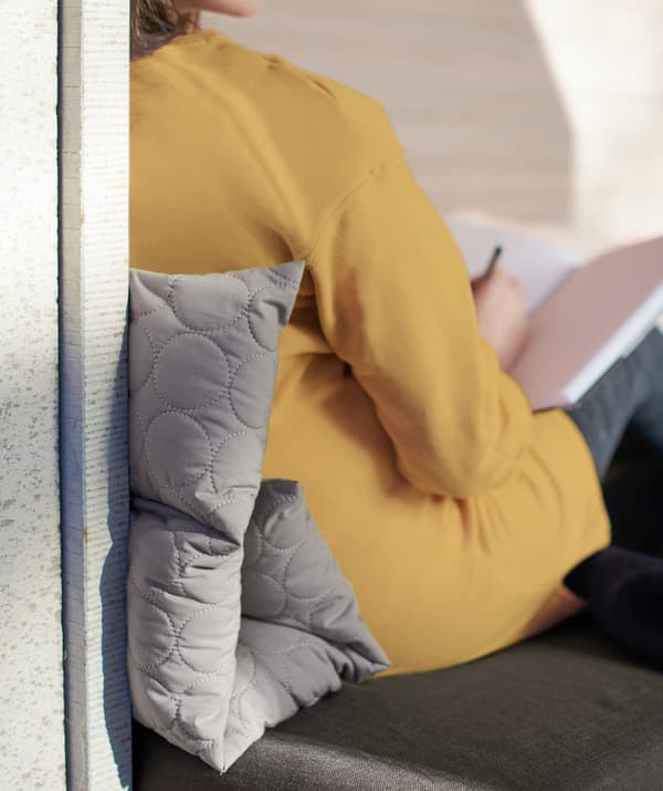 Femeie care își sprijină spatele, cu o pernă multifuncțională gri OMTÄNKSAM pliată între partea de jos a spatelui și peretele de care se sprijină.