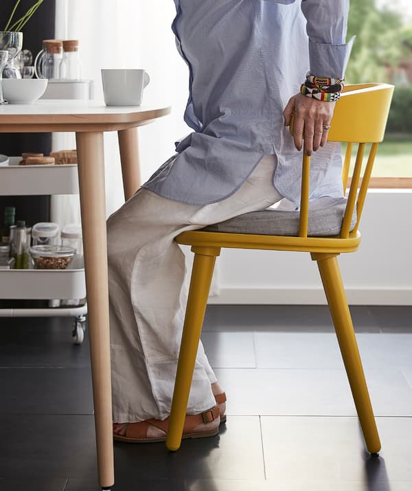 Femeie care se așază folosindu-și mâinile pentru a se sprijini de spătarul solid unui scaun galben OMTÄNKSAM.