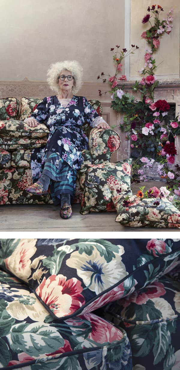 Femeie îmbrăcată cu o rochie cu flori pe o canapea cu motive florale lângă un șemineu decorat cu flori.