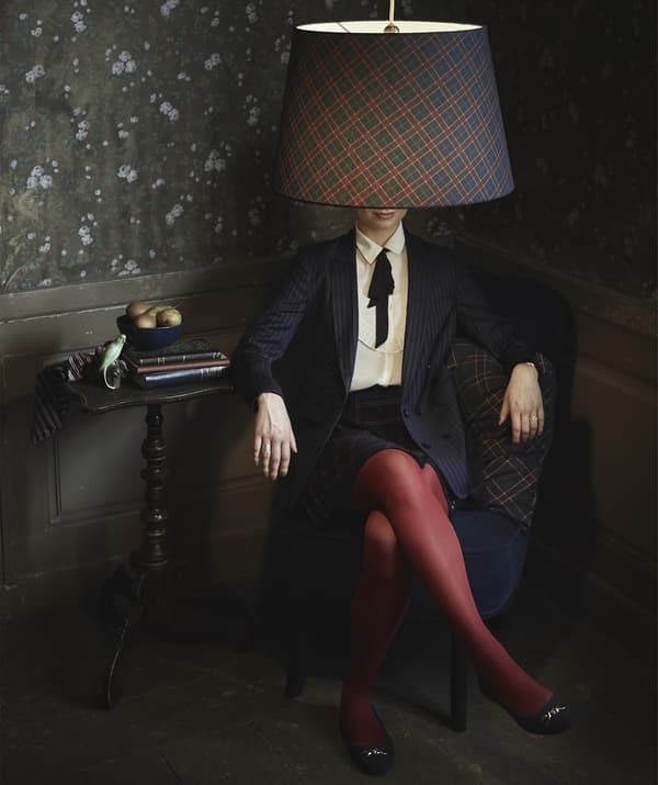 Femme assise dans un fauteuil, dans une pièce sombre, avec un abat-jour à motifs de carreaux à la place de la tête.