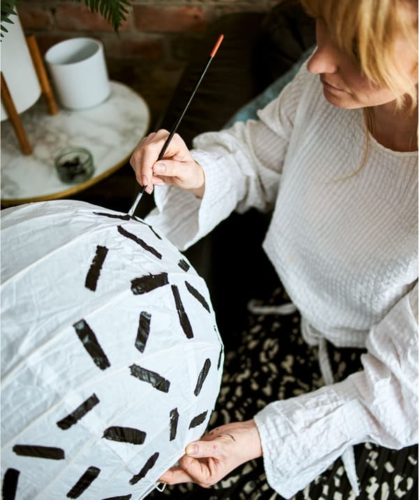 Femme aux cheveux blonds vêtue d'une chemise blanche en coton. Assise, elle peint des lignes noires épaisses sur un abat-jour en papier blanc.