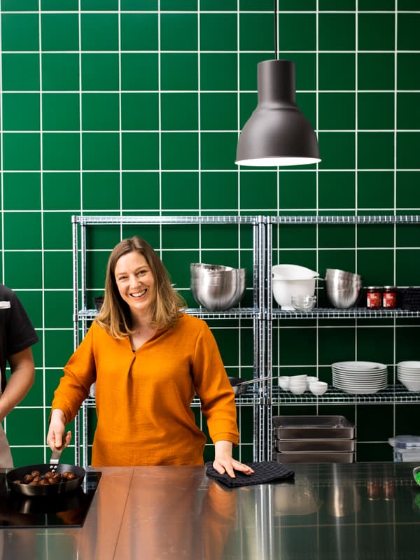 Femme debout dans une cuisine tout inox avec mur en carrelage vert tenant une poêle à frire pleine de boulettes végétales.