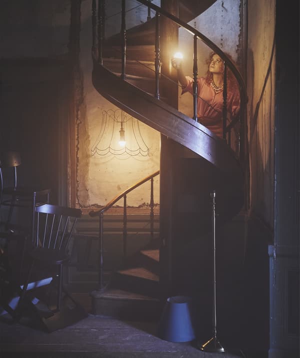 Femme éclairant ses pas dans l’obscurité avec une ampoule allumée, dans un escalier en colimaçon en bois.