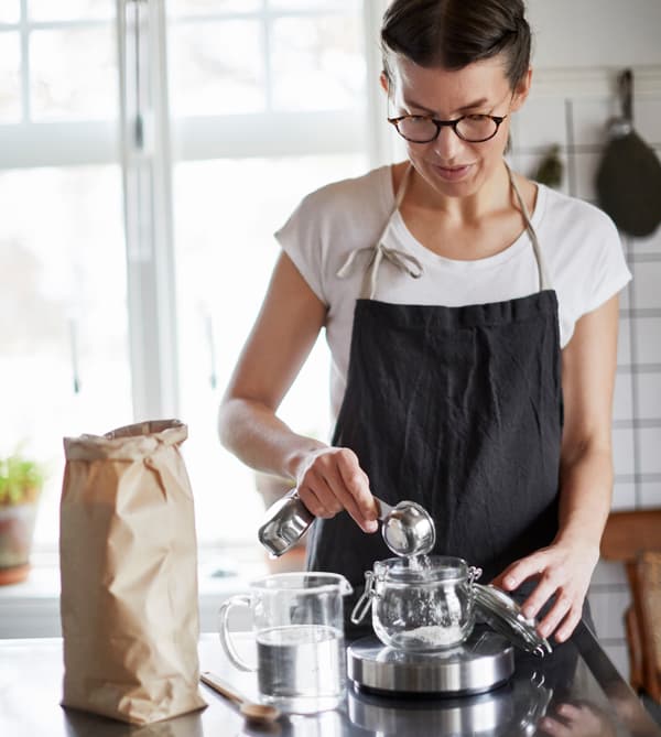 Femme en train de mesurer de la farine dans un bocal en verre, sur une balance de cuisine. 