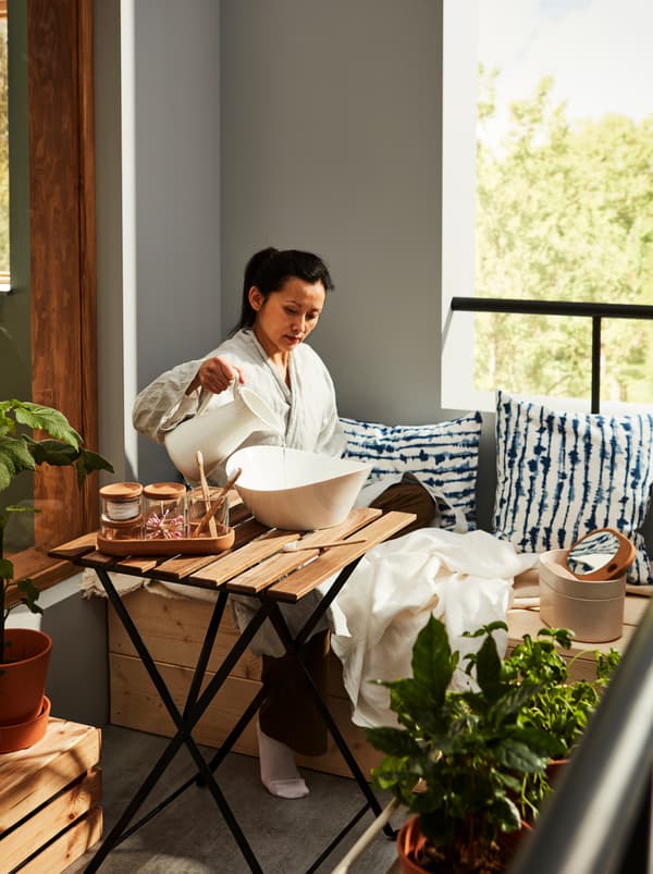 Femme vêtue d’un peignoir assise sur un balcon en train de verser de l’eau dans un saladier SKYN, avec des accessoires de toilette SAXBORGA posés à proximité.