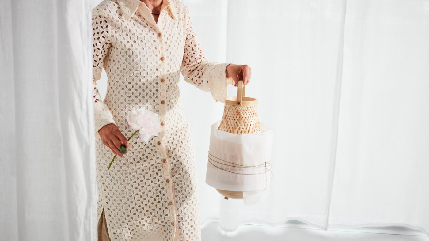 Femme vêtue d’une robe beige portant une lampe de table MISTERHULT en bambou tressé, partiellement enveloppée de papier.
