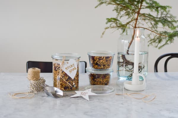 Festive scene with glasses filled with muesli bearing the inscription 'Merry Xmas', string and a metal scoop on a marble table. A burning candle and a small tree create a cosy atmosphere.