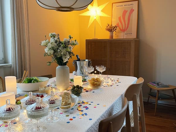 Festlich gedeckte, klassische Silvestertafel. Eine weiße Tischdecke, darauf zu sehen buntes Konfetti, eine Lichterkette und weiße LED Stumpenkerzen. EIne weiße STILREN VAse mit einem Blumenstrauß in weiß gehalten. SÄLLSKAPIG Sekt-und Dessertschalen schmücken den Tisch zusätzlich. 