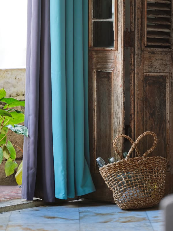 Fialové a tyrkysové zatemňovacie závesy zavesené na otvorených drevených dverách s pleteným košom položeným vedľa.