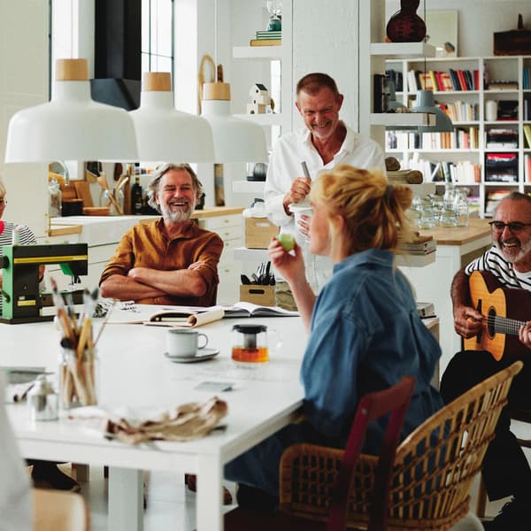 Fire mennesker sidder omkring et bord, drikker kaffe og snakker sammen, mens en mand spiller på guitar