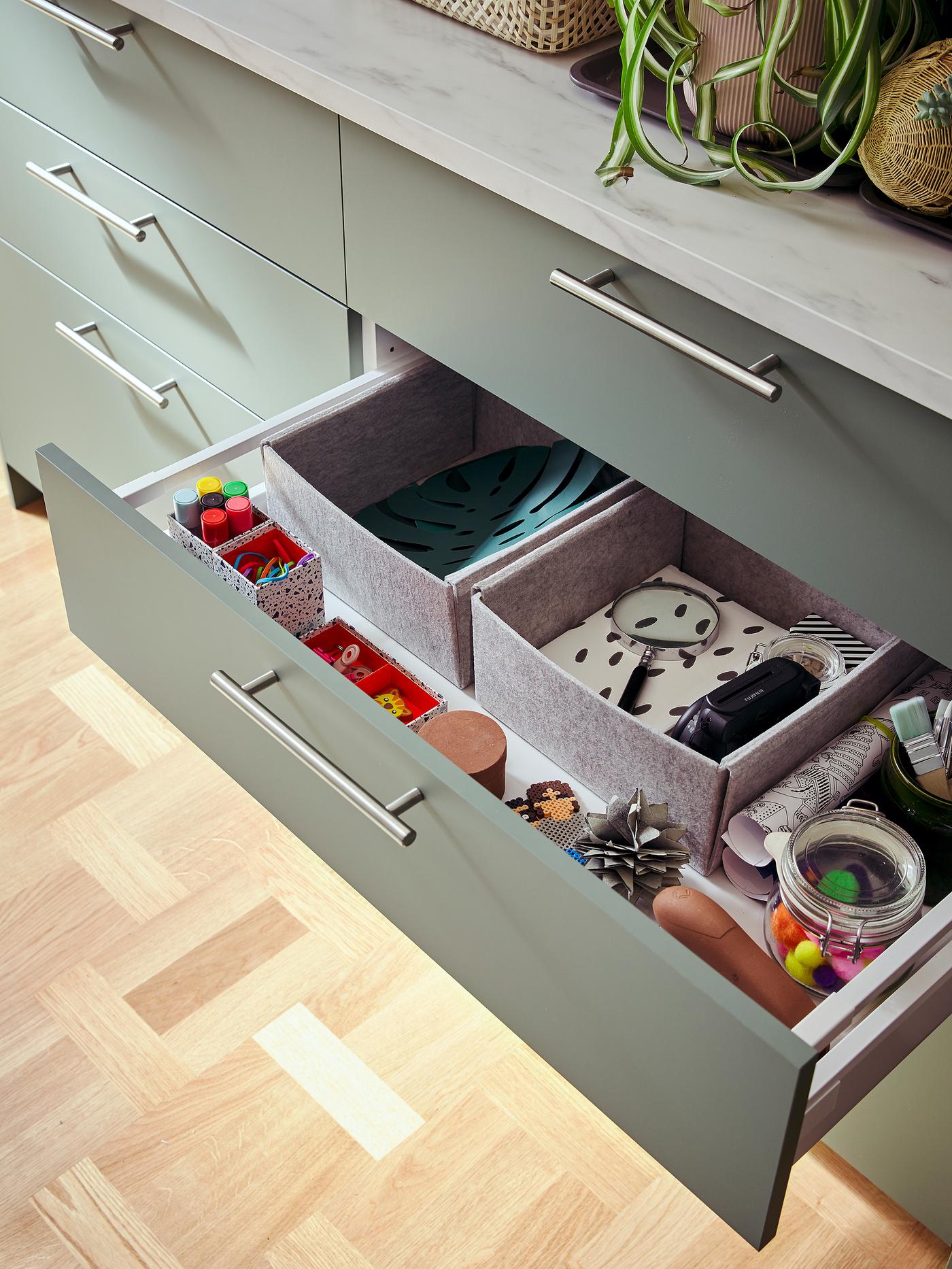 Five drawers in grey green below a light-grey mineral effect worktop. One drawer is open showing boxes, pens and more.