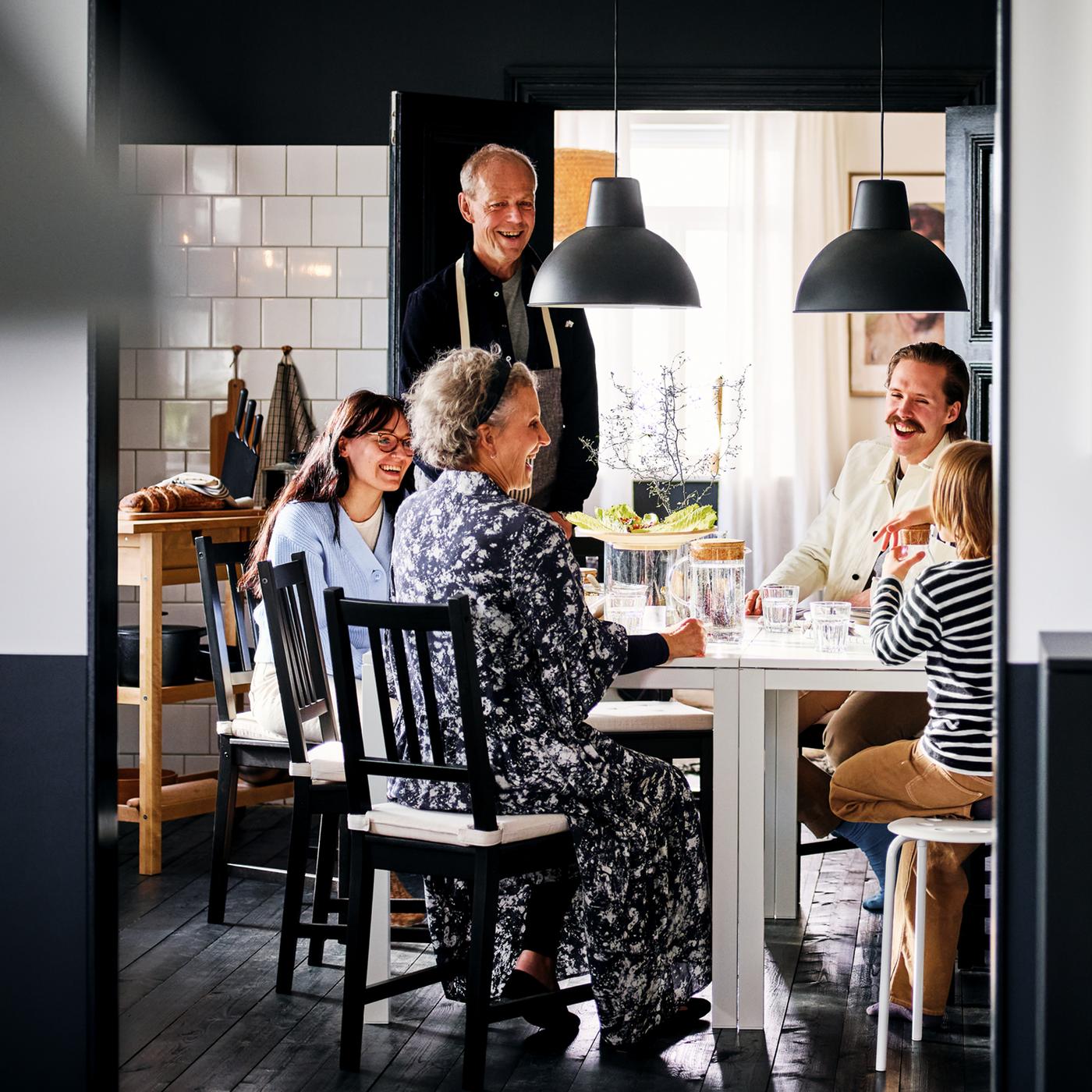 Five laughing people seated and standing around two white MELLTORP tables in a black and white dining area.