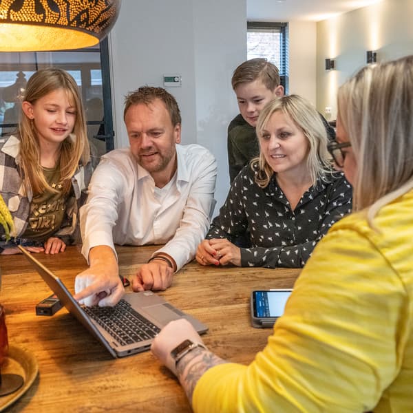 Five people gathered around wooden table, focused on laptop screen; an IKEA co-worker points at it under warm light in cozy indoor setting.