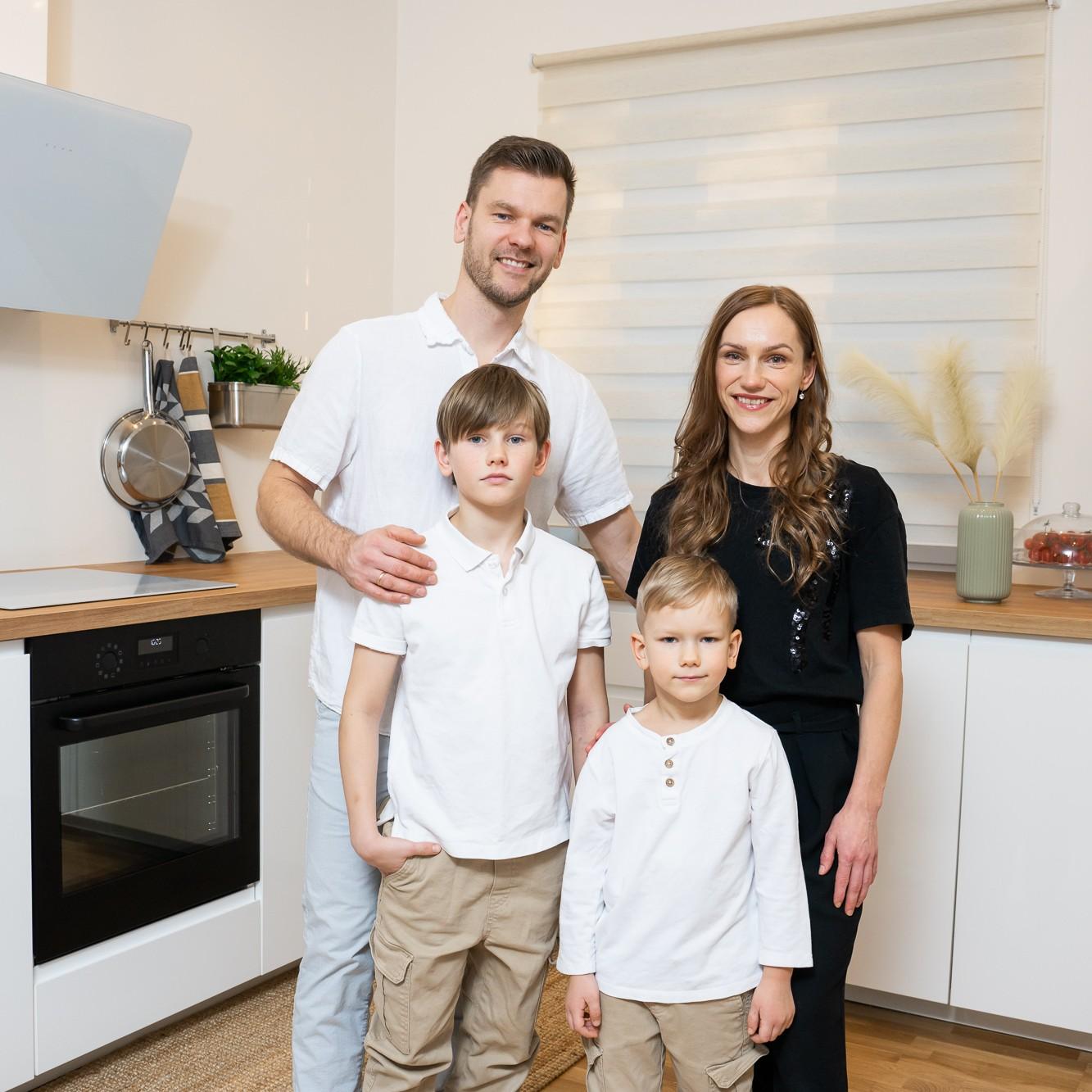 Five people standing in a bright, modern kitchen – two adults, two boys and an IKEA staff member. The family poses together with the designer next to white kitchen cabinets and a window with blinds.