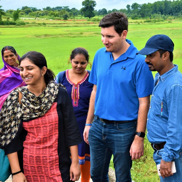 Five people standing outside in a field. Two are wearing blue shirts and the other three are wearing brightly coloured saris.