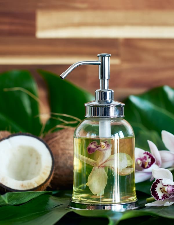 Flacon en verre rempli d’huile de massage et d'une fleur d’orchidée, avec en arrière-plan des plantes tropicales, des fleurs et des noix de coco. 