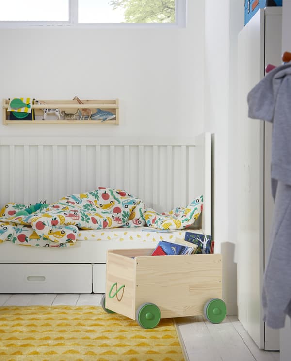FLISAT toy storage in pine with wheels and a handle in green is storing books, and it’s standing next to a white cot.