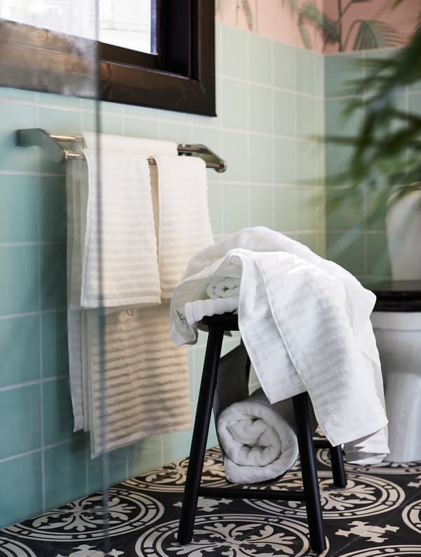 FLODALEN white bath towels that are placed on both a step stool and a towel rail in a pink/green/black bathroom.