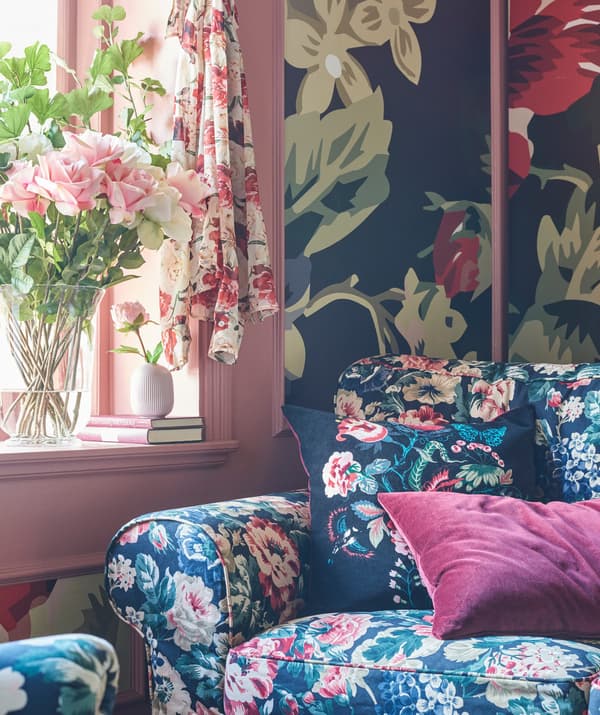 Floral patterned sofa with deep pink cushion in a room decorated with bold floral wallpaper and a pink window frame.