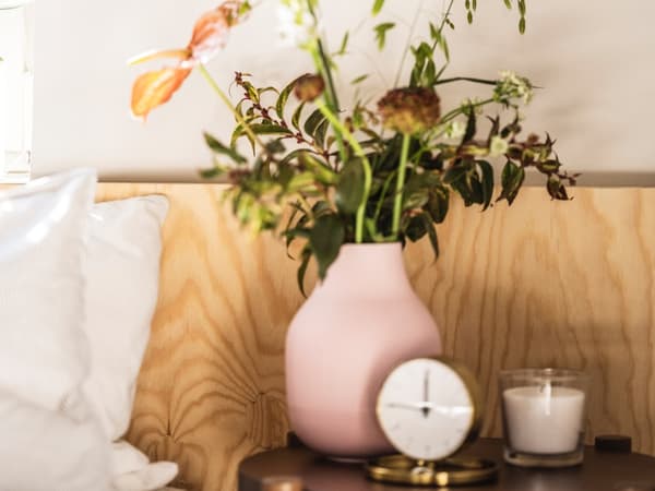 Florero de color rosado junto a un reloj y una vela, atras una pared de madera.