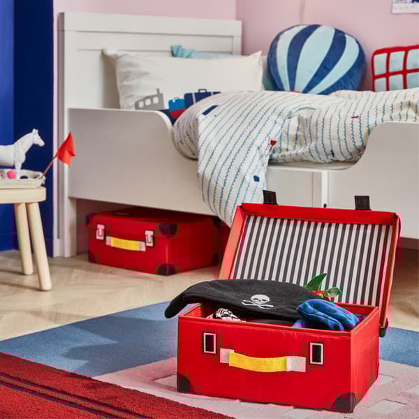 큰 배가 그려진 러그가 깔린 아이 방, 러그 위에 레드 색상의 FLYTTBAR 플뤼트바르 트렁크가 열려 있고 그 안에는 해적 옷이 들어 있는 모습