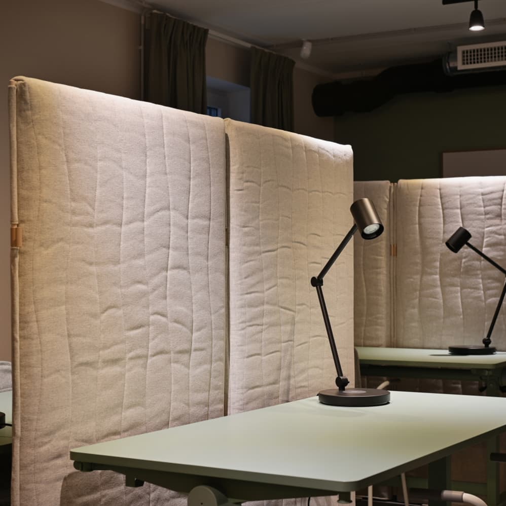 Focused workstations within a modern co-working space, separated by large acoustic divider panels. Adjustable desk lamps and muted tones create a calm and quiet environment designed for concentrated work.