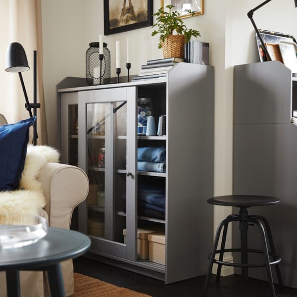 Folded jeans and boxes are stored inside a grey glass-door cabinet. A light beige sofa and a black stool are beside.