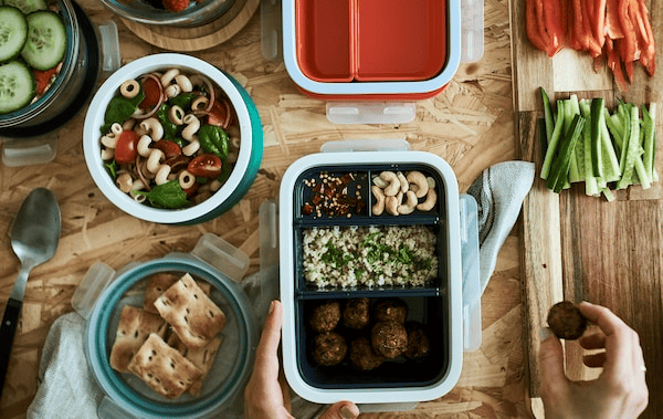 Food being packed into lunchboxes. One has compartments with different food, there’s pasta salad in a round plastic tub.