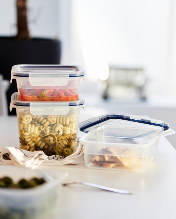 Food leftovers in three plastic food containers with lids, black/white tea towels and a fork on a white worktop.
