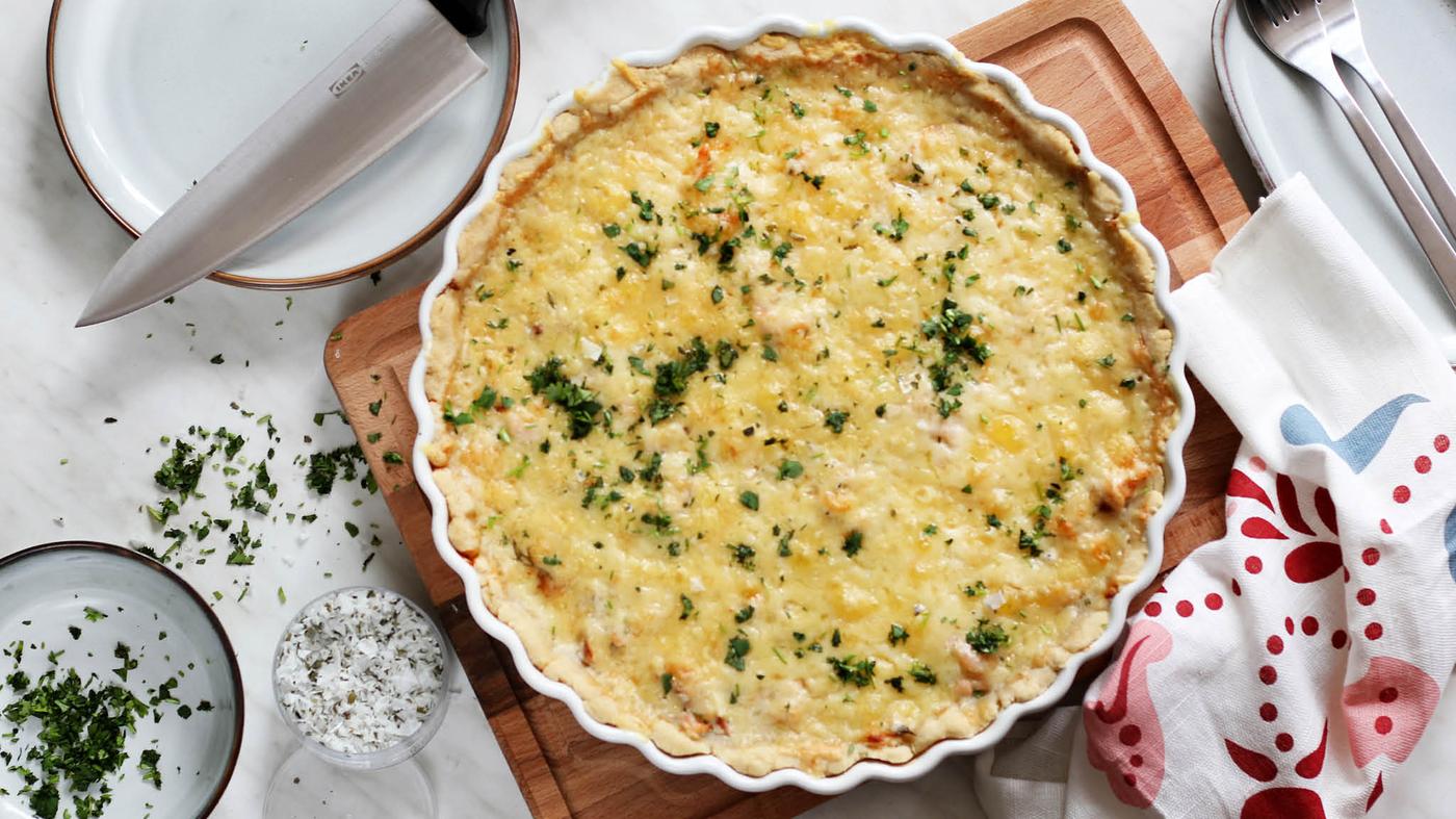 Forma na koláč VARDAGEN s krevetovo-lososovým slaným koláčem leží na prkénku PROPPMÄTT vedle utěrky a talířů GLADELIG.