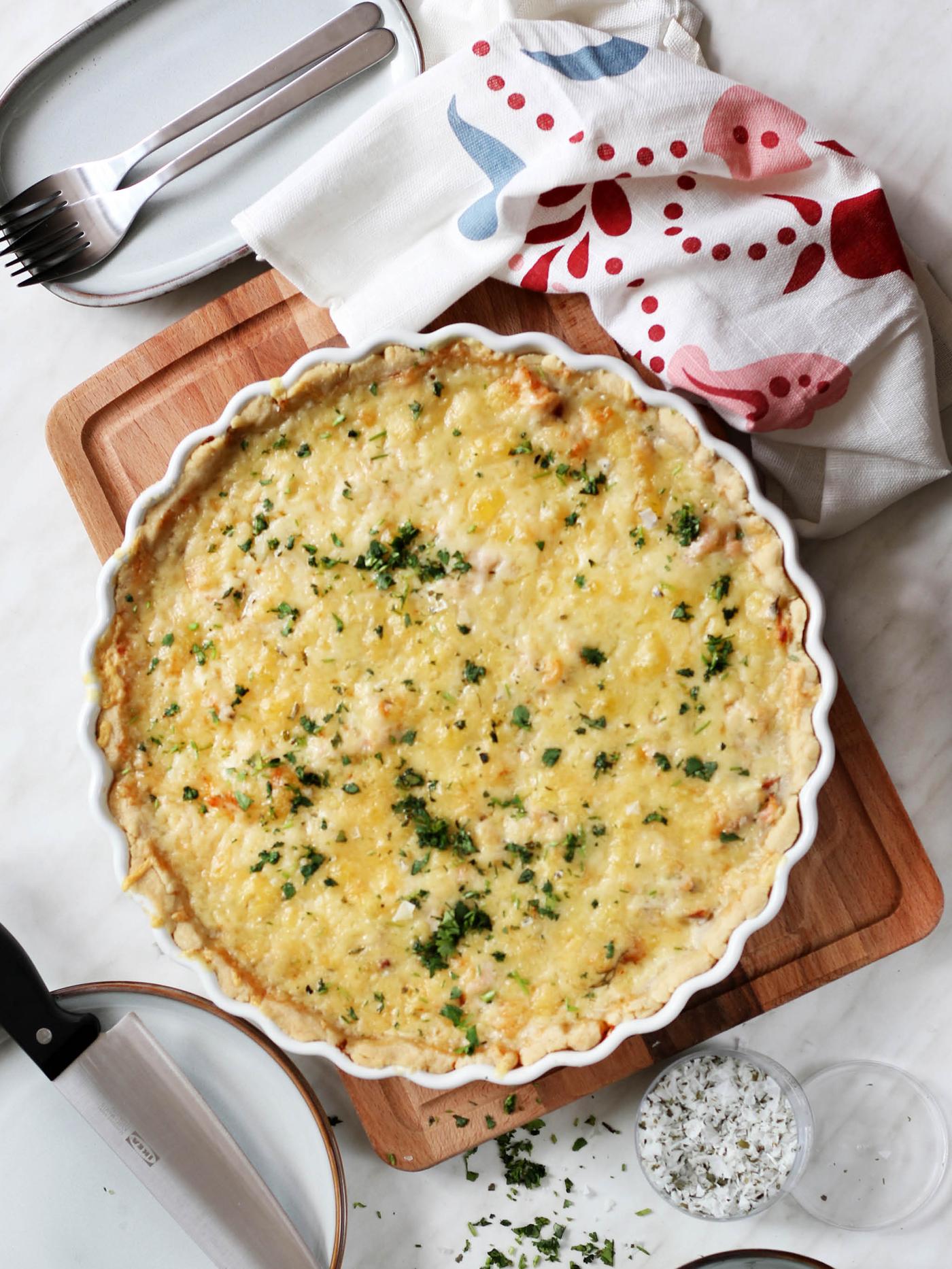 Forma na koláč VARDAGEN s krevetovo-lososovým slaným koláčem leží na prkénku PROPPMÄTT vedle utěrky a talířů GLADELIG.