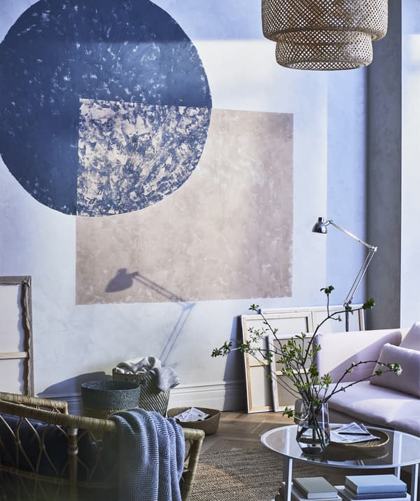 Formes géométriques peintes sur le mur d’un séjour devant un canapé rose et un fauteuil en rotin.