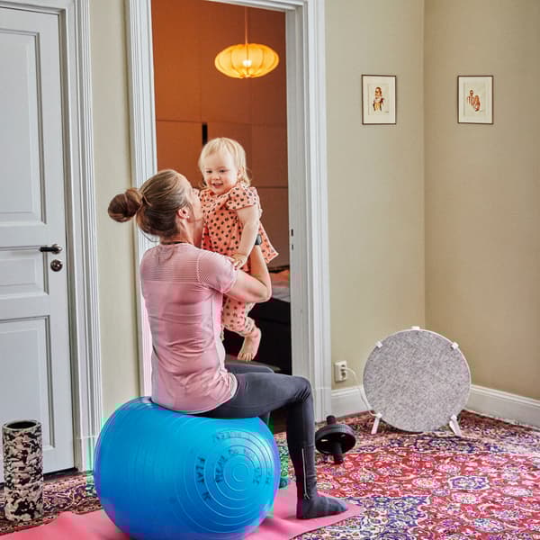 Foto på en mamma sittandes på en pilatesboll som leker med sin bebis. I bakgrunden står STARKVIND luftrenare från IKEA.