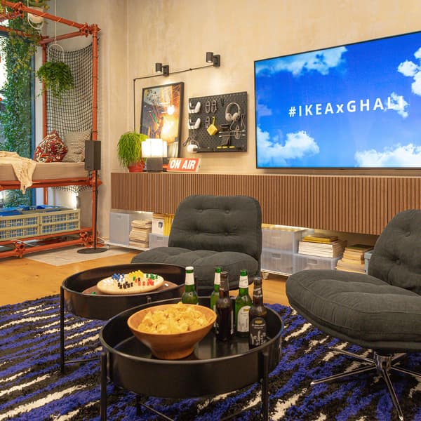 Foto panoramica del soggiorno di Casa Ghali con un tappeto, due tavolini bassi neri e due poltrone nere. Sulla parete c'è una grande tv accesa e dei dischi di Ghali.