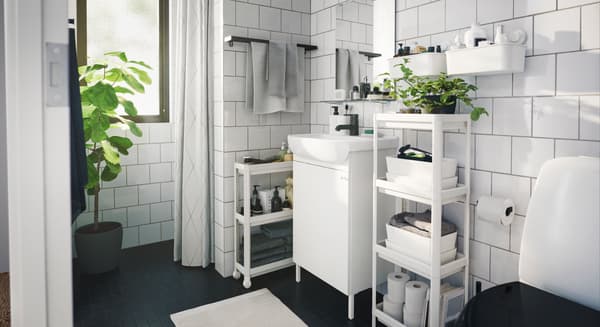 Fotografía lateral de un baño en colores fríos con muebles blancos y algunas plantas.
