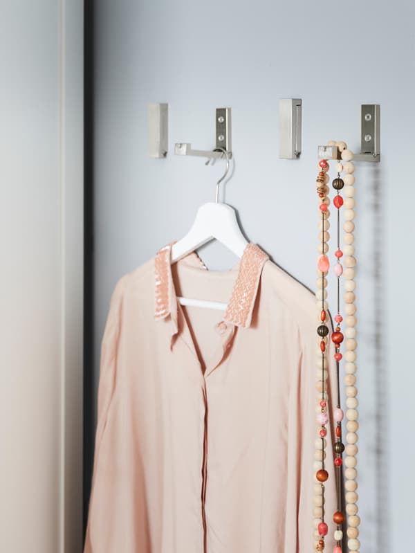 Four IKEA BJÄRNUM aluminium folding hooks on a wall next to a wardrobe, used to hang clothing and necklaces.