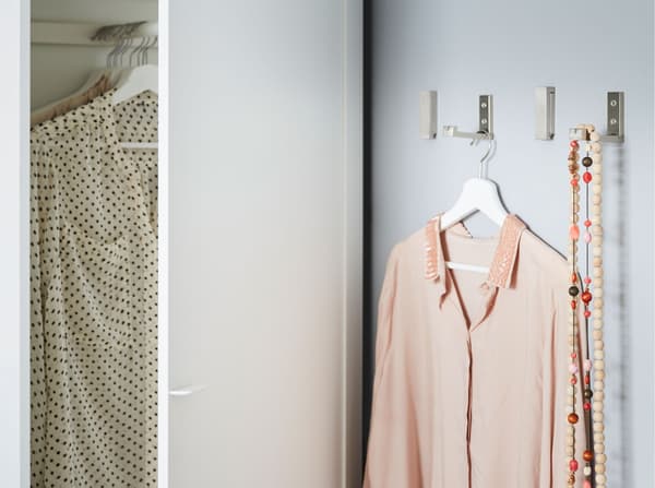 Four IKEA BJÄRNUM aluminium folding hooks on a wall next to a wardrobe, used to hang clothing and necklaces.
