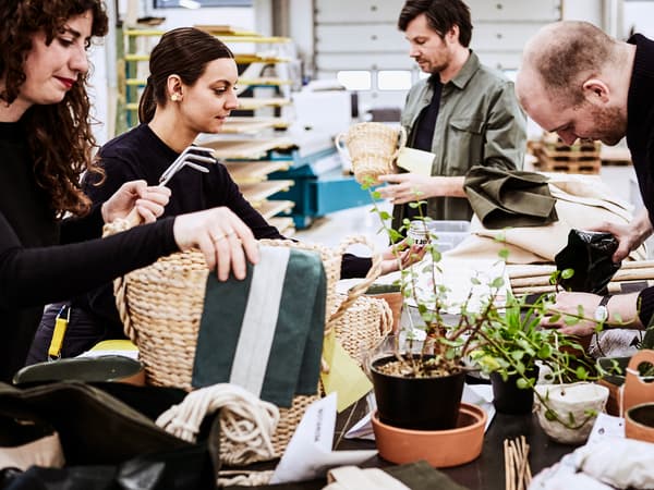 Four IKEA designers inspecting woven baskets around a large work top filled with bolts of folded material and pot plants.