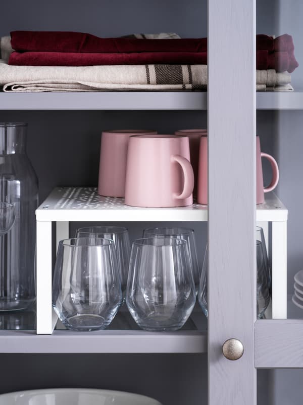 Four pink mugs on a white VARIERA shelf insert that stands above several drinking glasses on a grey cabinet shelf.