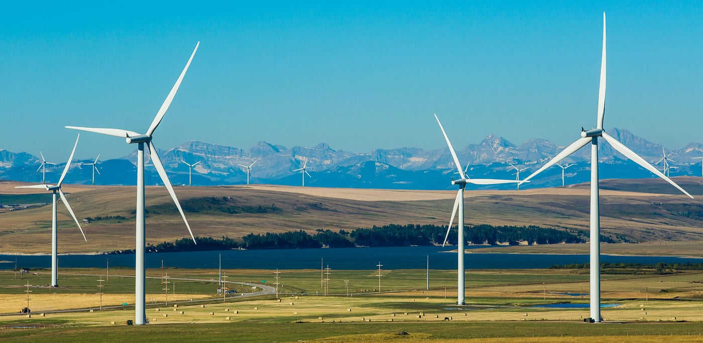 Four wind turbines in a green field with mountains and a blue sky in the background.
