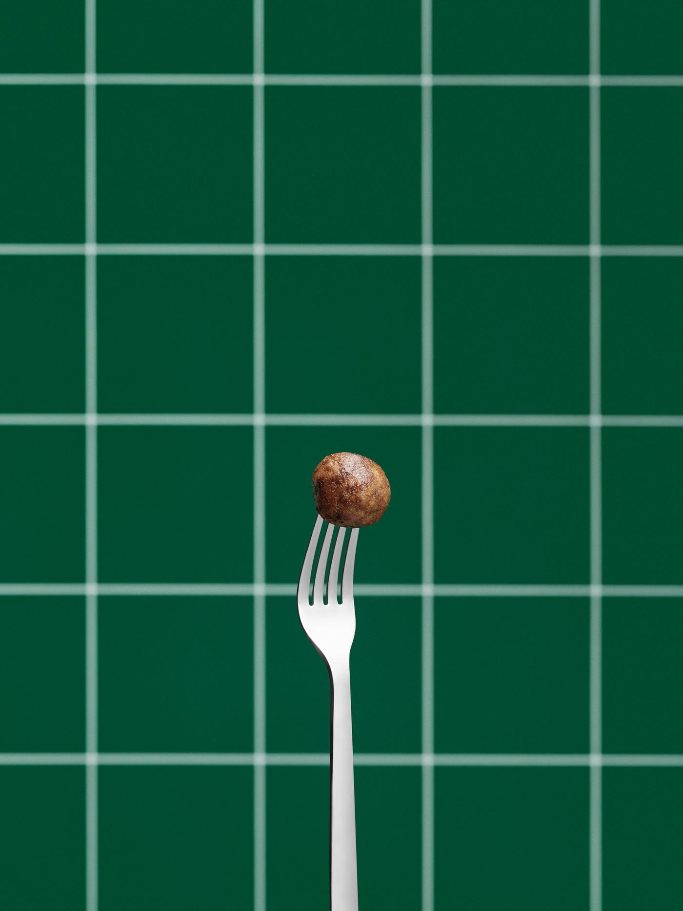 Fourchette en inox tenue verticalement, les dents piquées dans une boulette végétale devant un mur en carrelage vert.