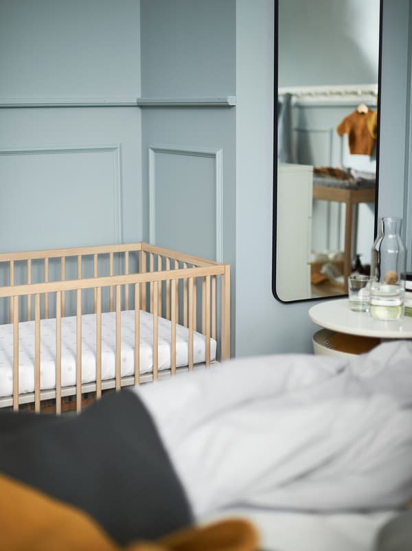 Fragment jasnoniebieskiej sypialni z białym stolikiem nocnym, lustrem i drewnianym łóżeczkiem dziecięcym SNIGLAR.