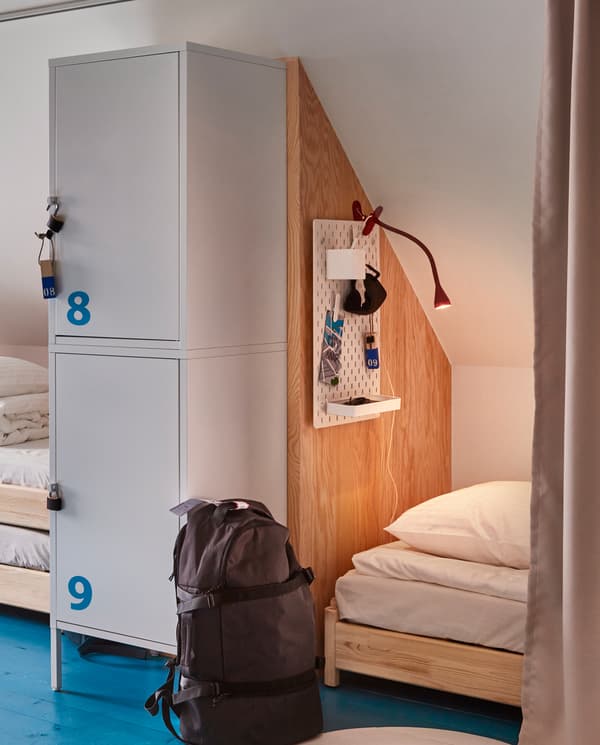 Fragmenty dwóch łóżek hostelowych oddzielonych ścianą i szafkami, przed którymi na podłodze stoi czarny plecak.